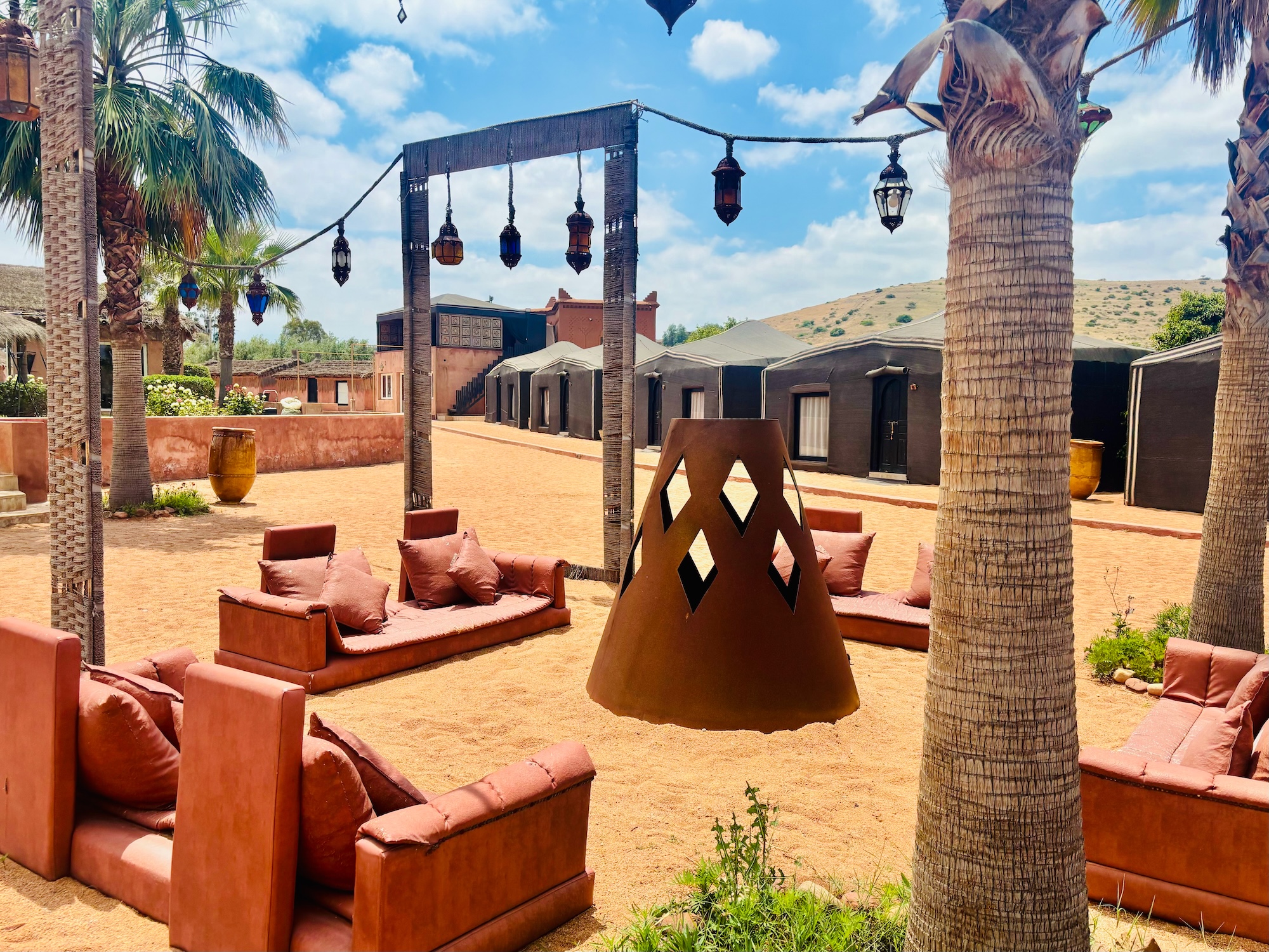 Surf Yoga Beer Does Morocco – An Experience in Novelty Beauty and Connection 2 - Marisa Markowitz, LCSW, CASAC, C-DBT - Marisa Markowitz, LCSW, CASAC, C-DBT Desert resort courtyard with orange couches, palm trees, and hanging lanterns on a sunny day.