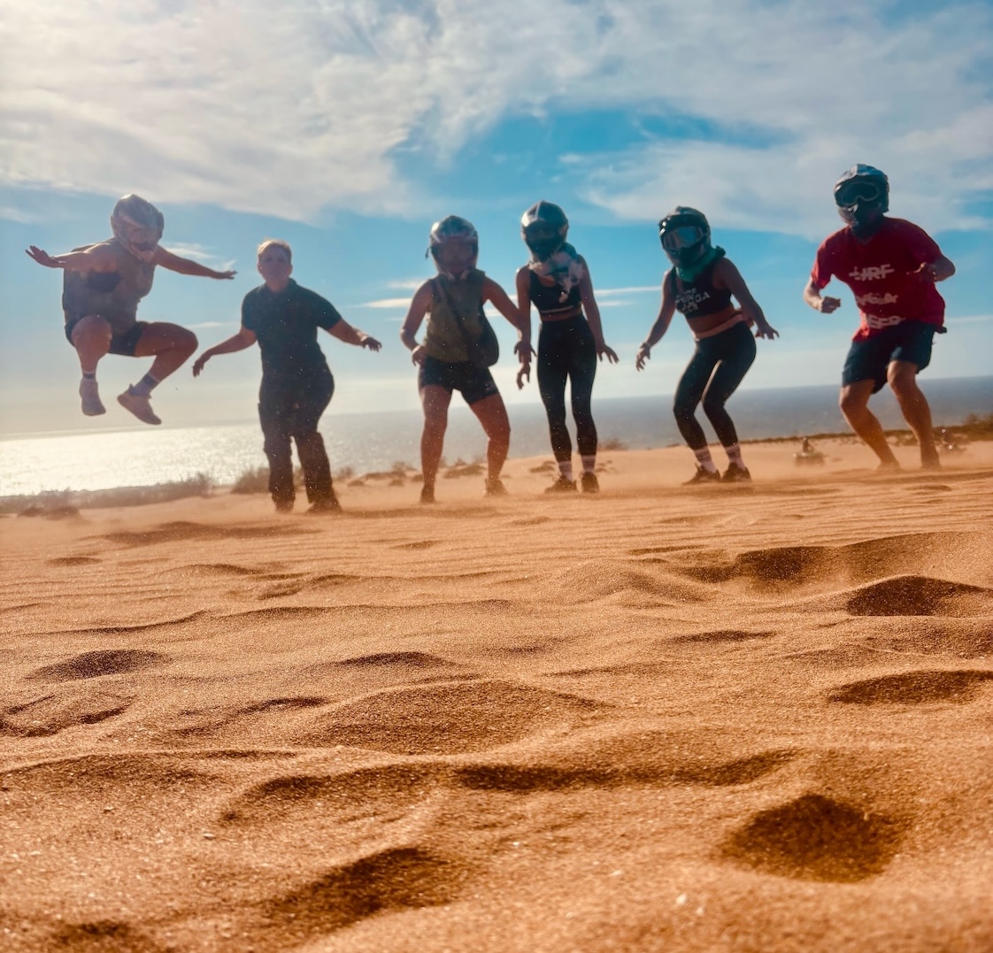 Six people wearing protective gear jump in unison on a sunny beach, with the ocean and blue sky in the background.
